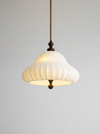 Close angled view of the lit white pleated shade on this Clérachic pendant, with its brass rod and walnut detail on a grey backdrop.