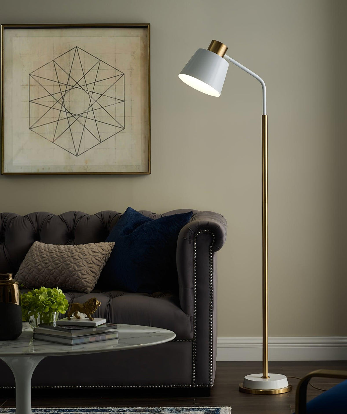 Clérachic floor lamp in white and gold behind a grey leather chesterfield, with a geometric framed print on a pale wall.