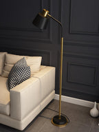 A Clérachic floor lamp with a black shade on a bent arm, beside a cream sofa and a white vase on a grey tiled floor.