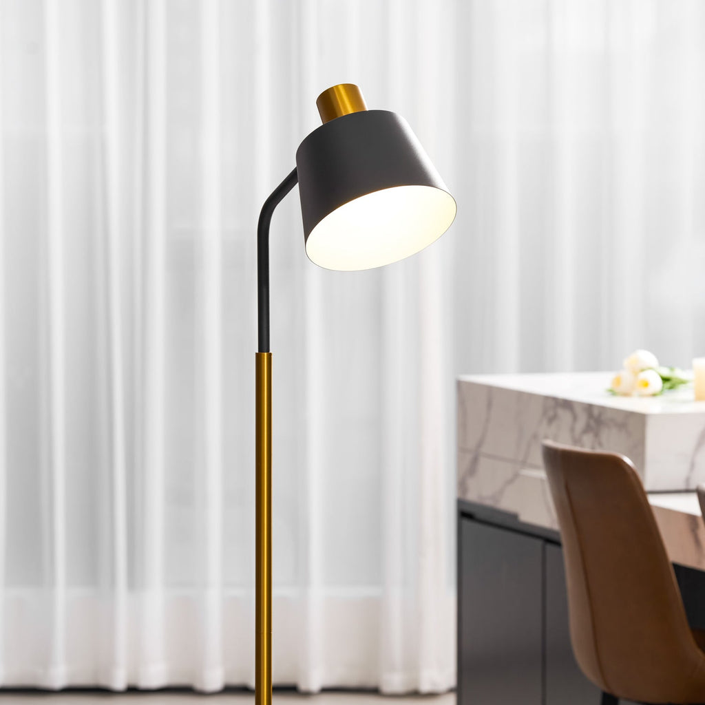 Close view of the black shade, gold collar and curved arm of this Clérachic floor lamp, lit before sheer white curtains.
