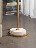Close view of the white round base with a gold rim on this Clérachic floor lamp, standing on grey floor tiles by a stack of books.