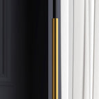 Detail of the slim gold stem of this Clérachic floor lamp, with a dark wall on one side and a white curtain on the other.