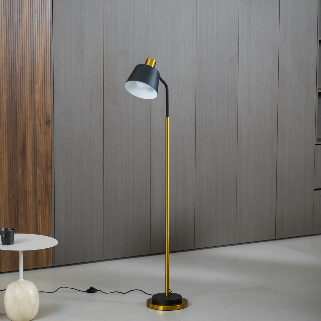 A Clérachic floor lamp in black and gold against grey wood-panelled walls, with a round white side table holding a dark cup.
