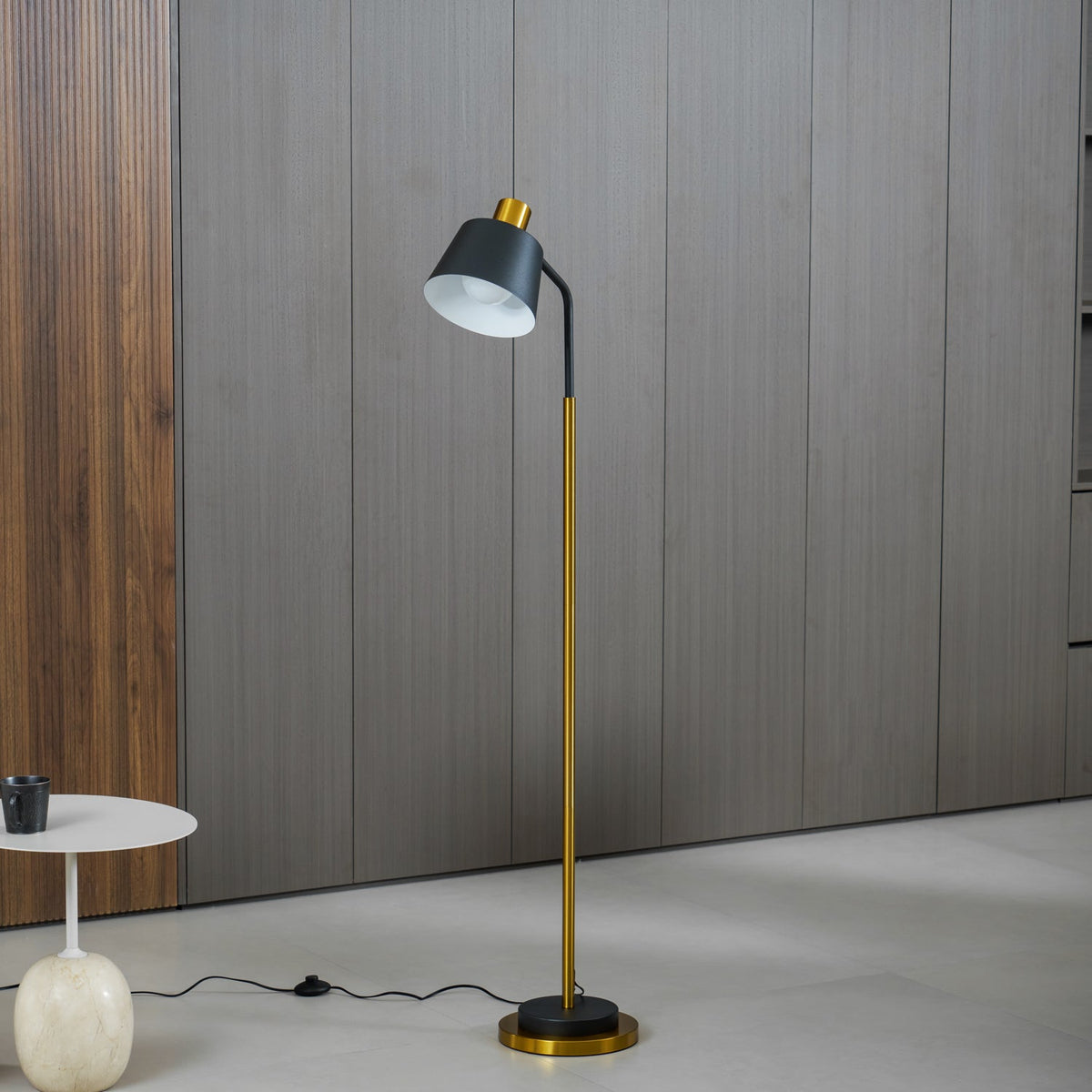 A Clérachic floor lamp in black and gold against grey wood-panelled walls, with a round white side table holding a dark cup.