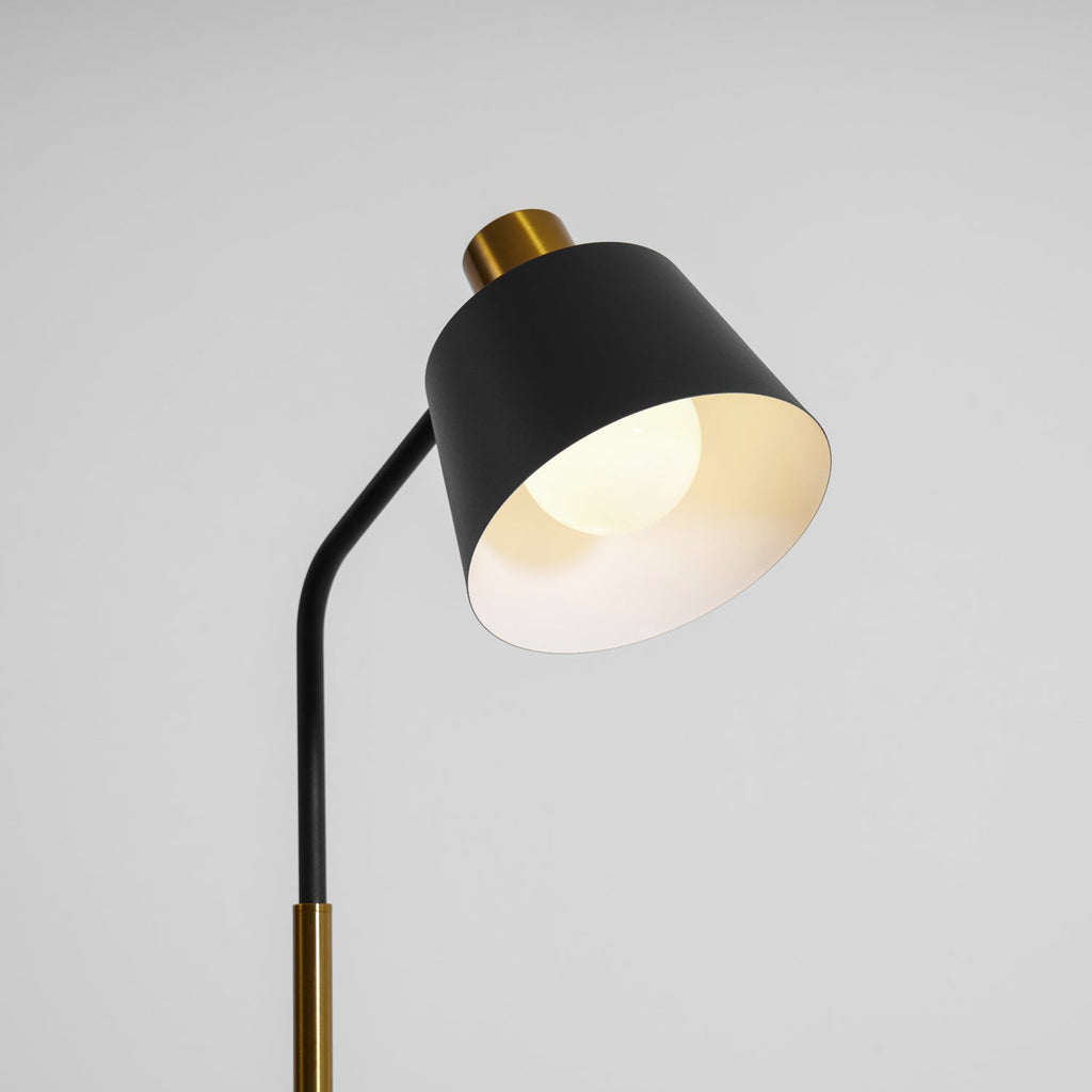 Black shade of this Clérachic floor lamp tilted down with the bulb glowing warm inside, against a light grey wall.
