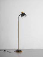 Clérachic floor lamp in black and gold on a grey studio floor, its shade tilted up and the cord switch lying beside the base.