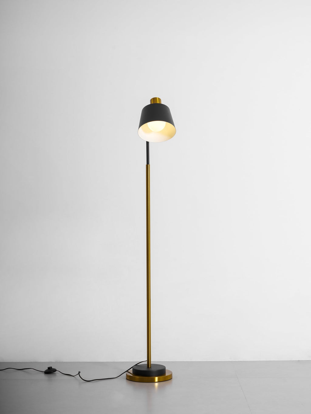 Clérachic floor lamp with its black shade turned upward, the lit inside glowing, on a grey studio floor.