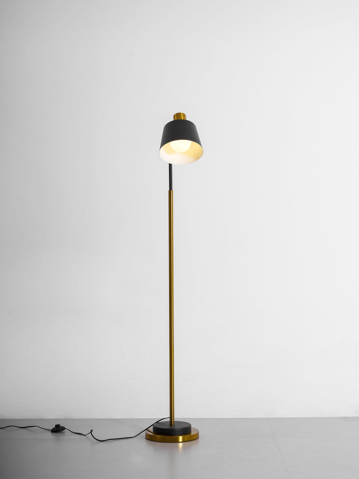 Clérachic floor lamp with its black shade turned upward, the lit inside glowing, on a grey studio floor.