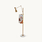 Gold Clérachic floor lamp with a cone shade on an angled arm, a framed picture on its pole and a round white marble base, on a plain backdrop.