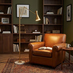 A Clérachic gold floor lamp leans its cone shade over a tan leather armchair, with dark wood bookshelves and a patterned rug behind it.