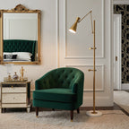 Lit cone shade on a Clérachic gold floor lamp beside a green tufted armchair, with a gold-framed mirror and a mirrored side table.