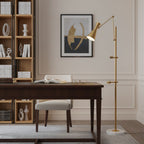 A Clérachic gold floor lamp stands by a dark wood desk with its shade lit toward an abstract print, with open shelves behind.