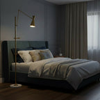 The gold cone shade of this Clérachic floor lamp glows in a dim bedroom, beside a bed with grey and cream bedding and a heavy curtain.