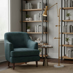 A Clérachic gold floor lamp with a marble base stands by a teal armchair and a metal shelving unit with books and small plants.