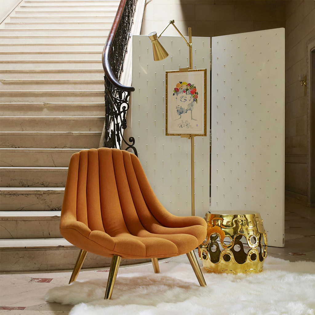 A Clérachic gold floor lamp holding a small framed portrait, beside an orange velvet armchair and a gold side stool at the foot of a staircase.