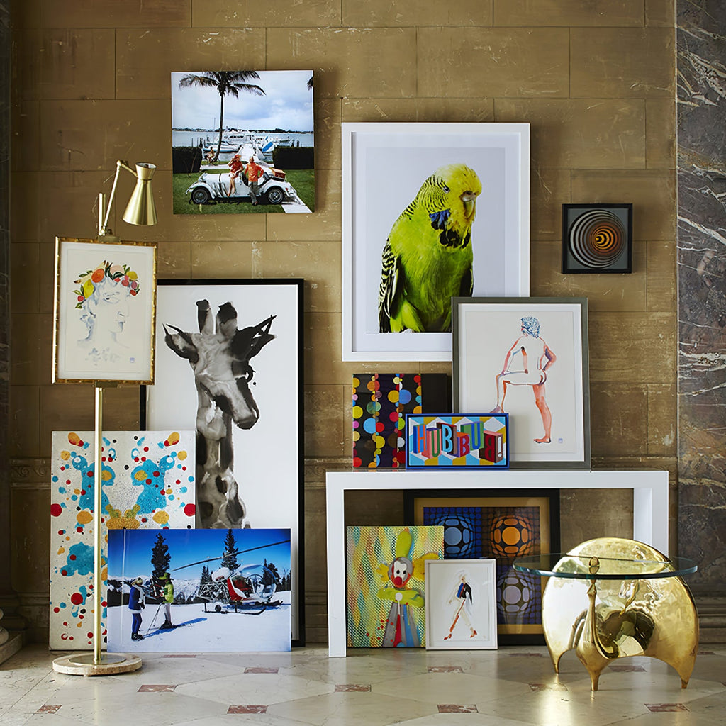 A Clérachic gold floor lamp with a framed print among a crowded gallery of colourful artworks, with a gold side table and a stone wall.