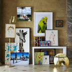 A Clérachic gold floor lamp with a framed print among a crowded gallery of colourful artworks, with a gold side table and a stone wall.