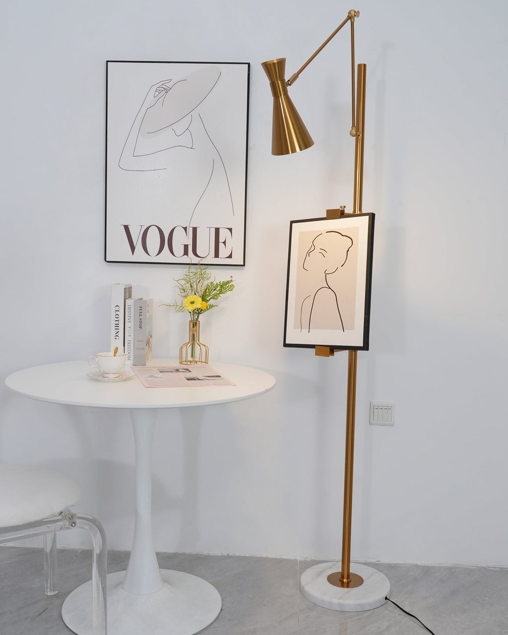 Gold Clérachic floor lamp with a framed line drawing on its pole, beside a round white table with a small vase of flowers.