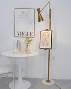 Gold Clérachic floor lamp with a framed line drawing on its pole, beside a round white table with a small vase of flowers.