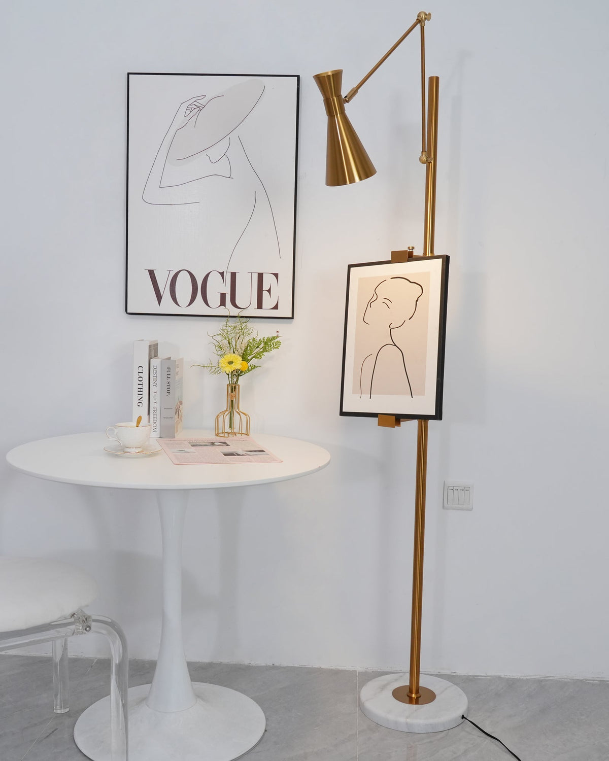 Gold Clérachic floor lamp with a framed line drawing on its pole, beside a round white table with a small vase of flowers.
