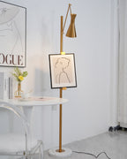 Close view of the pole of this Clérachic gold floor lamp with a framed line drawing, beside a round white table and a clear chair.
