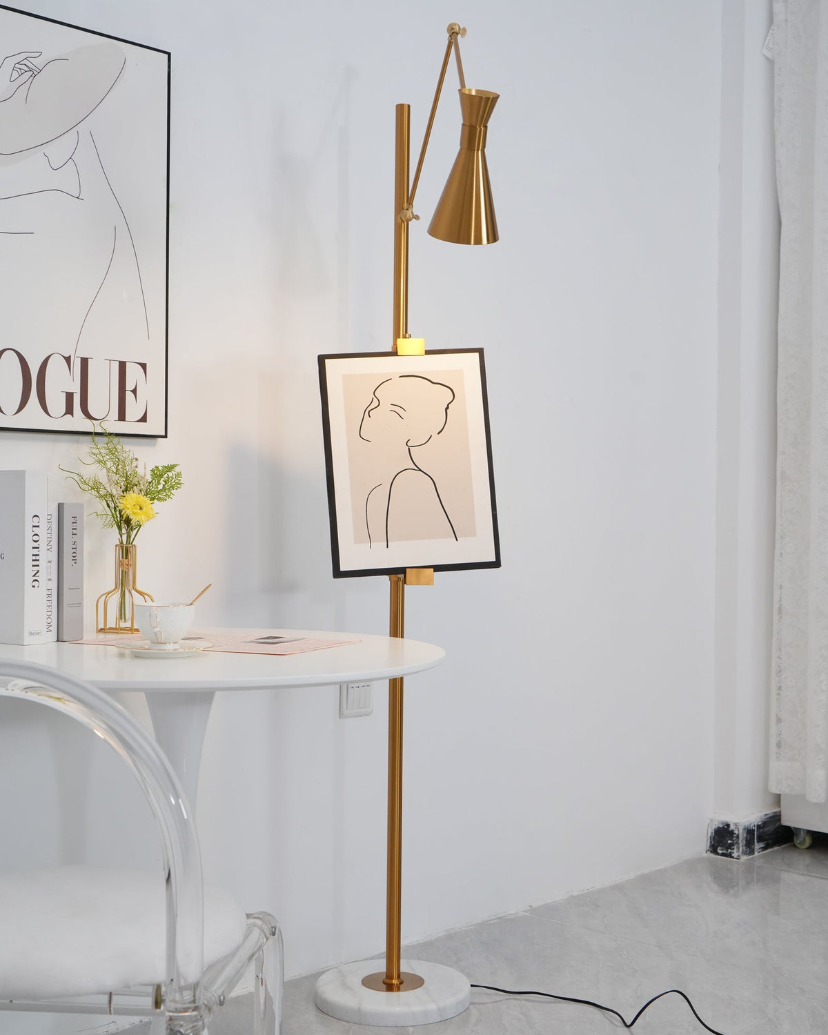 Close view of the pole of this Clérachic gold floor lamp with a framed line drawing, beside a round white table and a clear chair.