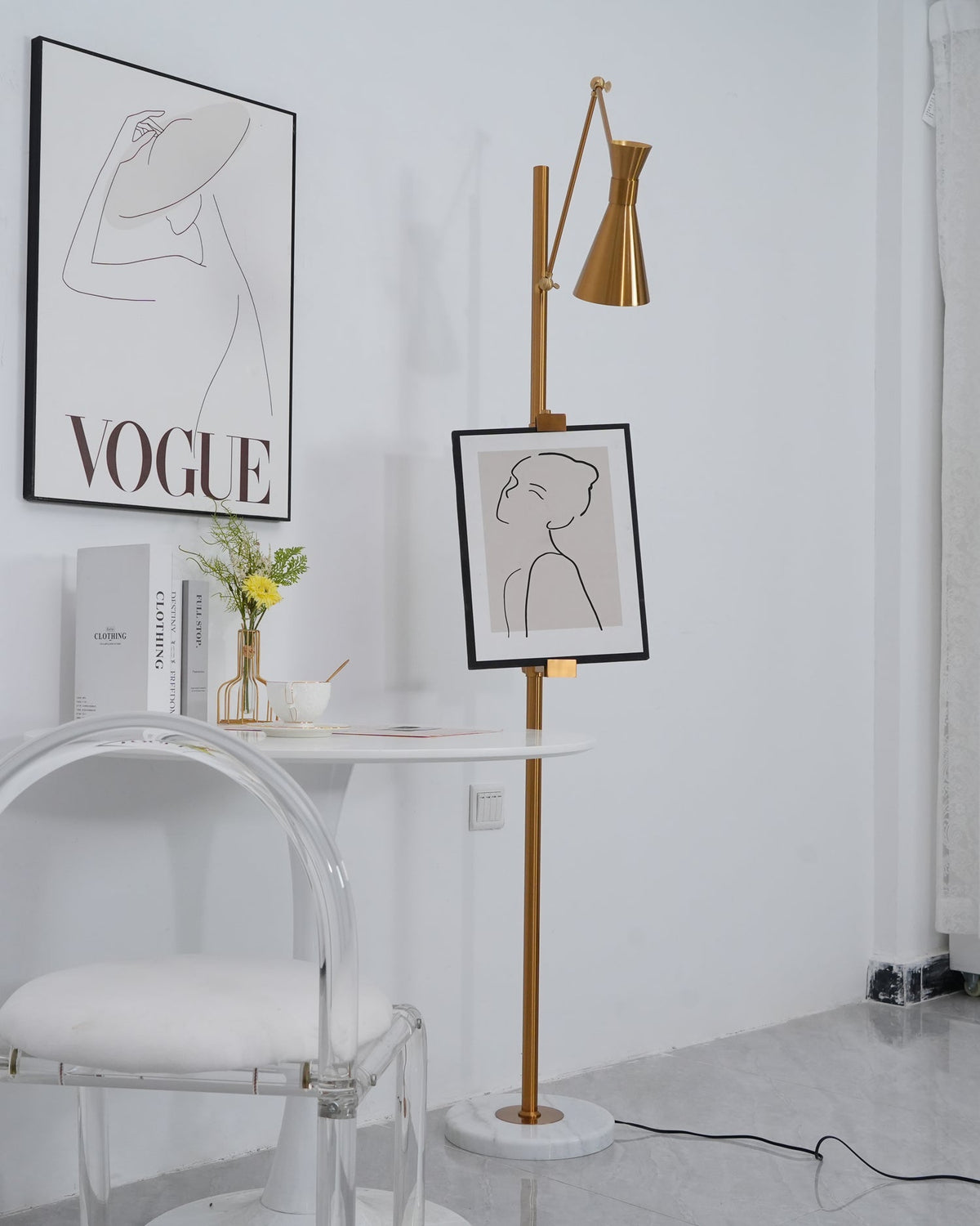 Cone shade and framed drawing on a Clérachic gold floor lamp, next to a white table with a vase of yellow flowers and a clear chair.