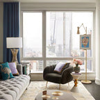A Clérachic gold floor lamp with a small framed picture stands by a large window with a city view, near a grey tufted sofa and a dark armchair.
