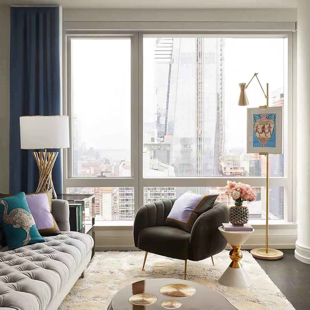 A Clérachic gold floor lamp with a small framed picture stands by a large window with a city view, near a grey tufted sofa and a dark armchair.