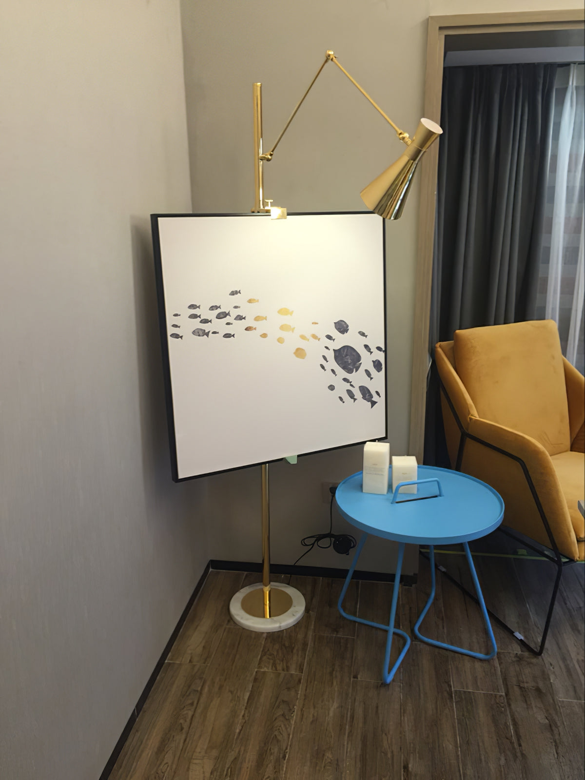 Large framed picture mounted on the pole of a Clérachic gold floor lamp in a corner, with a blue side table and a mustard armchair.