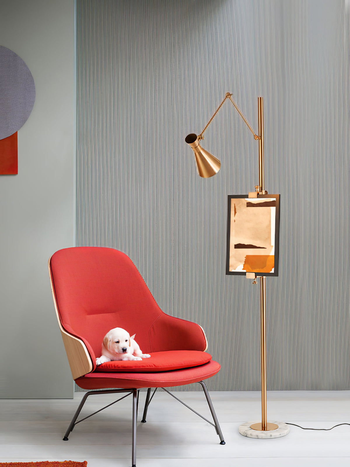 A Clérachic gold floor lamp with a framed copper-toned print, beside a red lounge chair with a small white puppy on the seat.