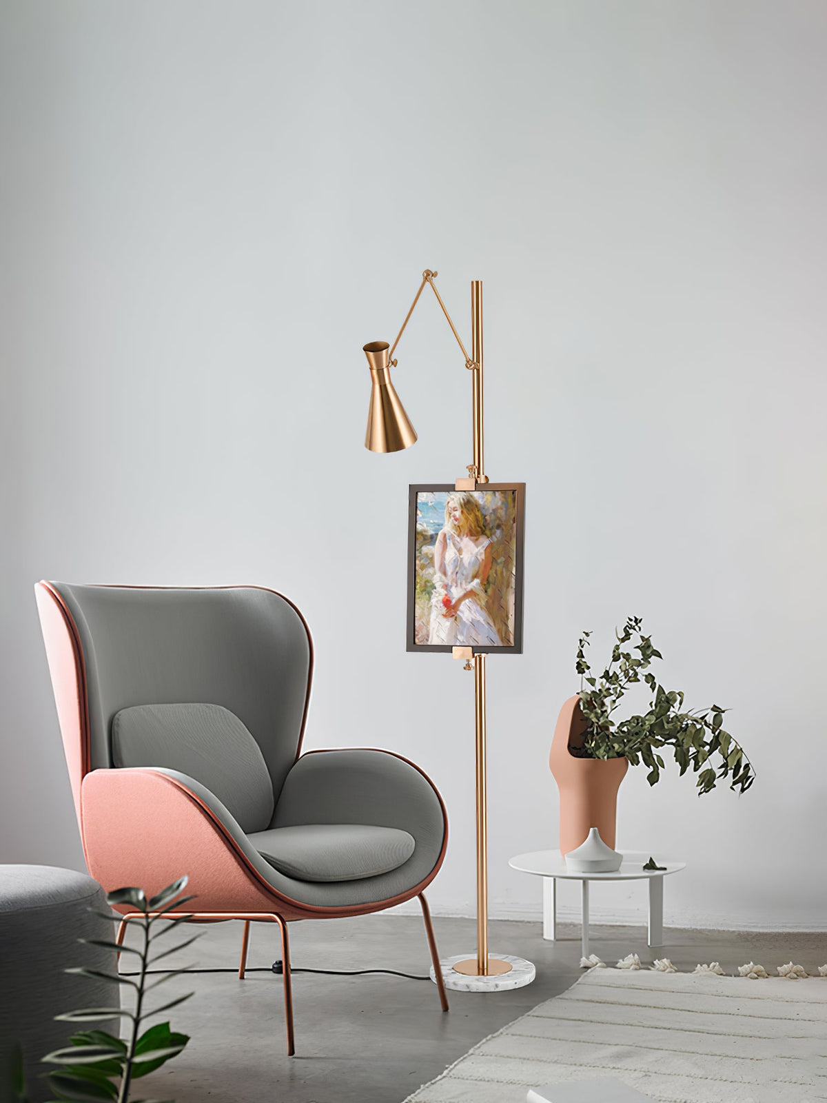Clérachic gold floor lamp with a small framed portrait on its pole, beside a grey and pink armchair and a tall plant.