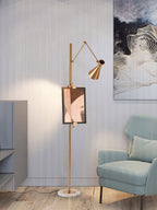 A Clérachic gold floor lamp with a framed print on its pole stands by a pale blue armchair, with a wall-art panel behind.