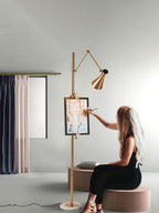A person sits in front of this Clérachic gold floor lamp and touches a framed picture clipped to its pole, in a grey room with curtains.