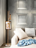 Clérachic gold floor lamp with a framed picture in a room with grey panelled walls, near a cream sofa with a pale blue throw.