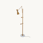 Gold Clérachic floor lamp with an arc arm, cone shade and two small clamp arms on the pole, above a white marble base on a cream backdrop.
