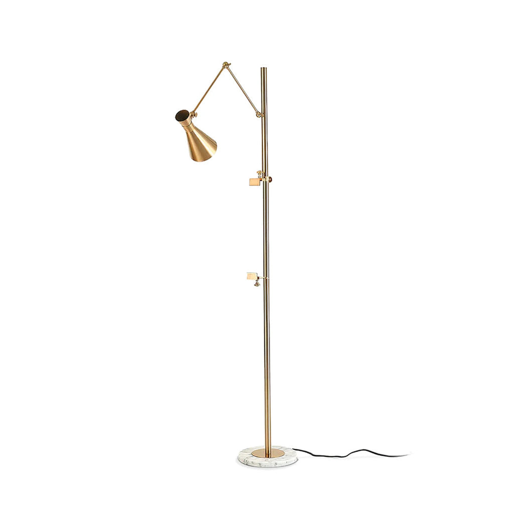 Slim gold Clérachic floor lamp with a cone shade, two clamp arms and a black cord trailing from a marble base, on a white backdrop.