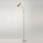 A Clérachic gold floor lamp stands alone against a soft grey wall, with a cone shade at the top and a marble base on the floor.