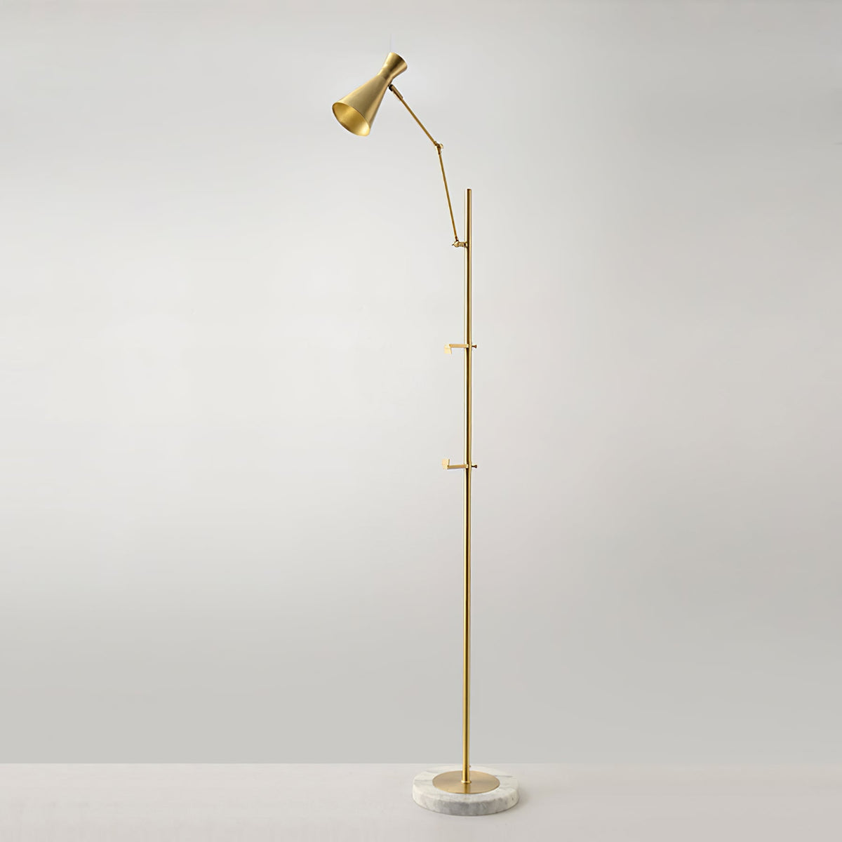 A Clérachic gold floor lamp stands alone against a soft grey wall, with a cone shade at the top and a marble base on the floor.