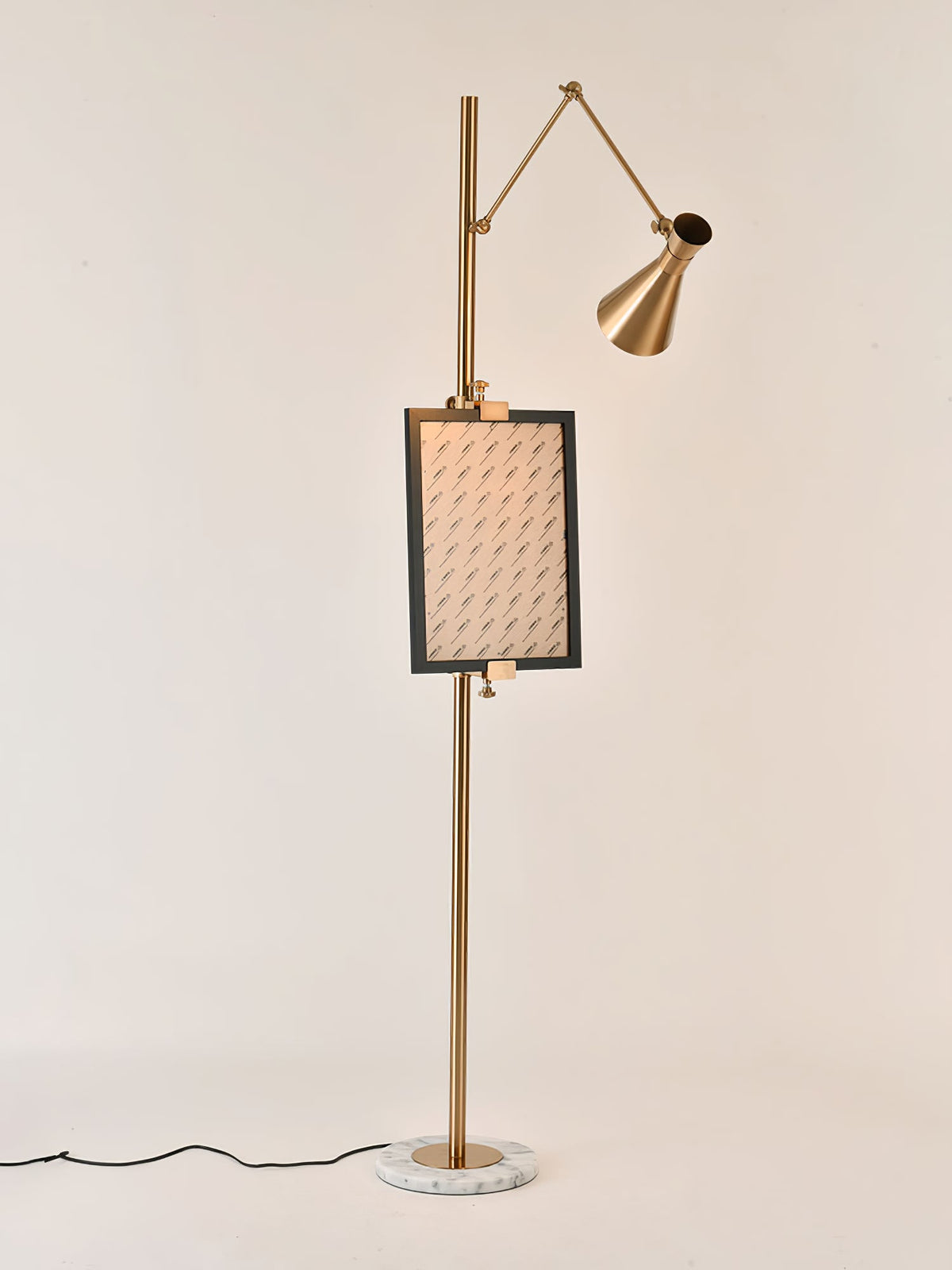 Gold Clérachic floor lamp with a framed picture clipped to the pole and a black cord running from a white marble base, on a peach backdrop.