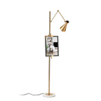 A Clérachic gold floor lamp with a framed picture on its pole and a black cord trailing from the marble base, on a white backdrop.