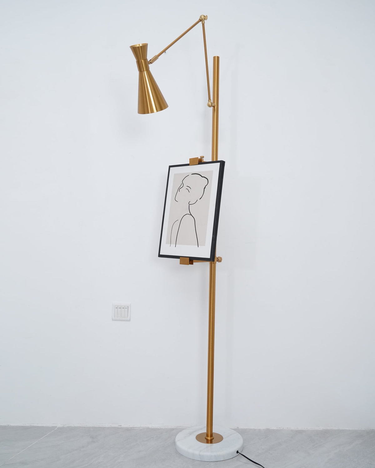 A tall Clérachic gold floor lamp with a framed line drawing on the pole, in front of a plain white wall with a power socket.