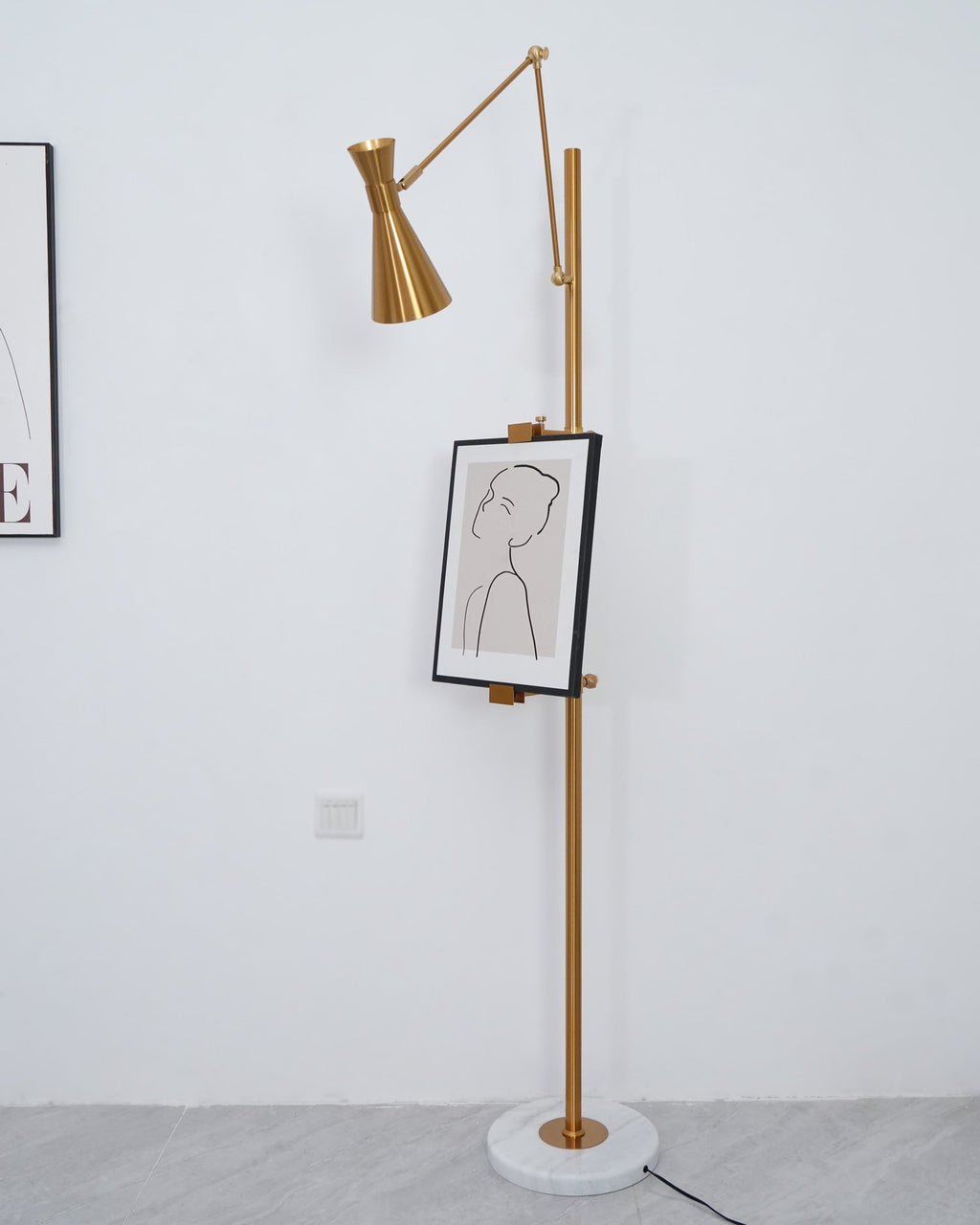 A Clérachic gold floor lamp with a framed line drawing, standing against a white wall with the edge of a print at the left.