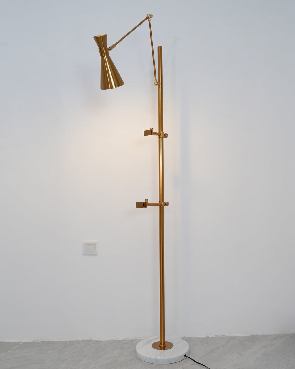 A lit gold cone shade on a Clérachic floor lamp throws a soft glow on a white wall, with two clamp arms on the pole below.