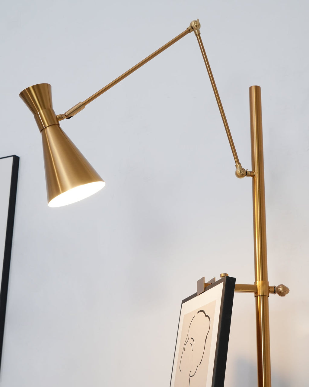 Lit gold cone shade on a Clérachic floor lamp, angled down above a framed line drawing clipped to its pole.