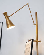 Lit gold cone shade on a Clérachic floor lamp, angled down above a framed line drawing clipped to its pole.