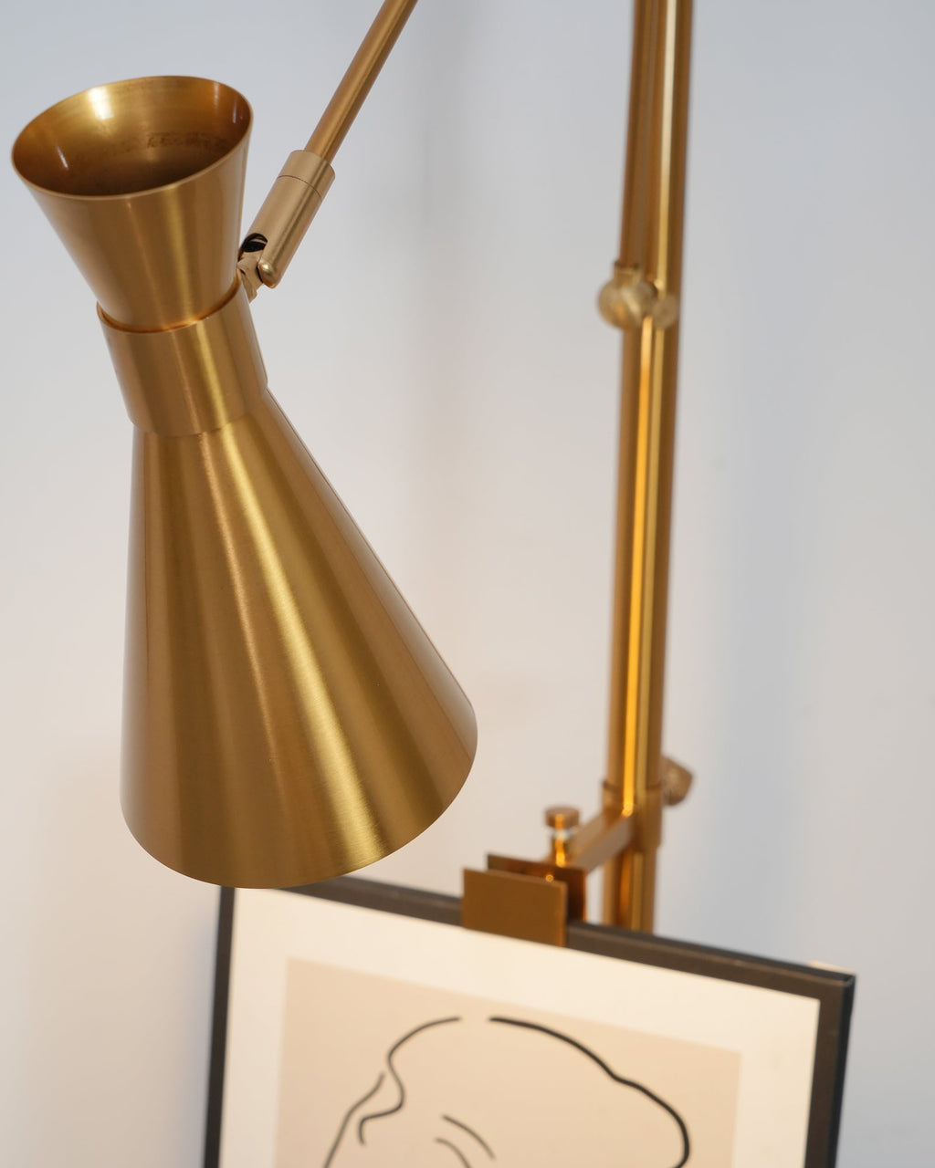 Close view of the gold cone shade of a Clérachic floor lamp above a framed drawing, with the arm joint just behind.