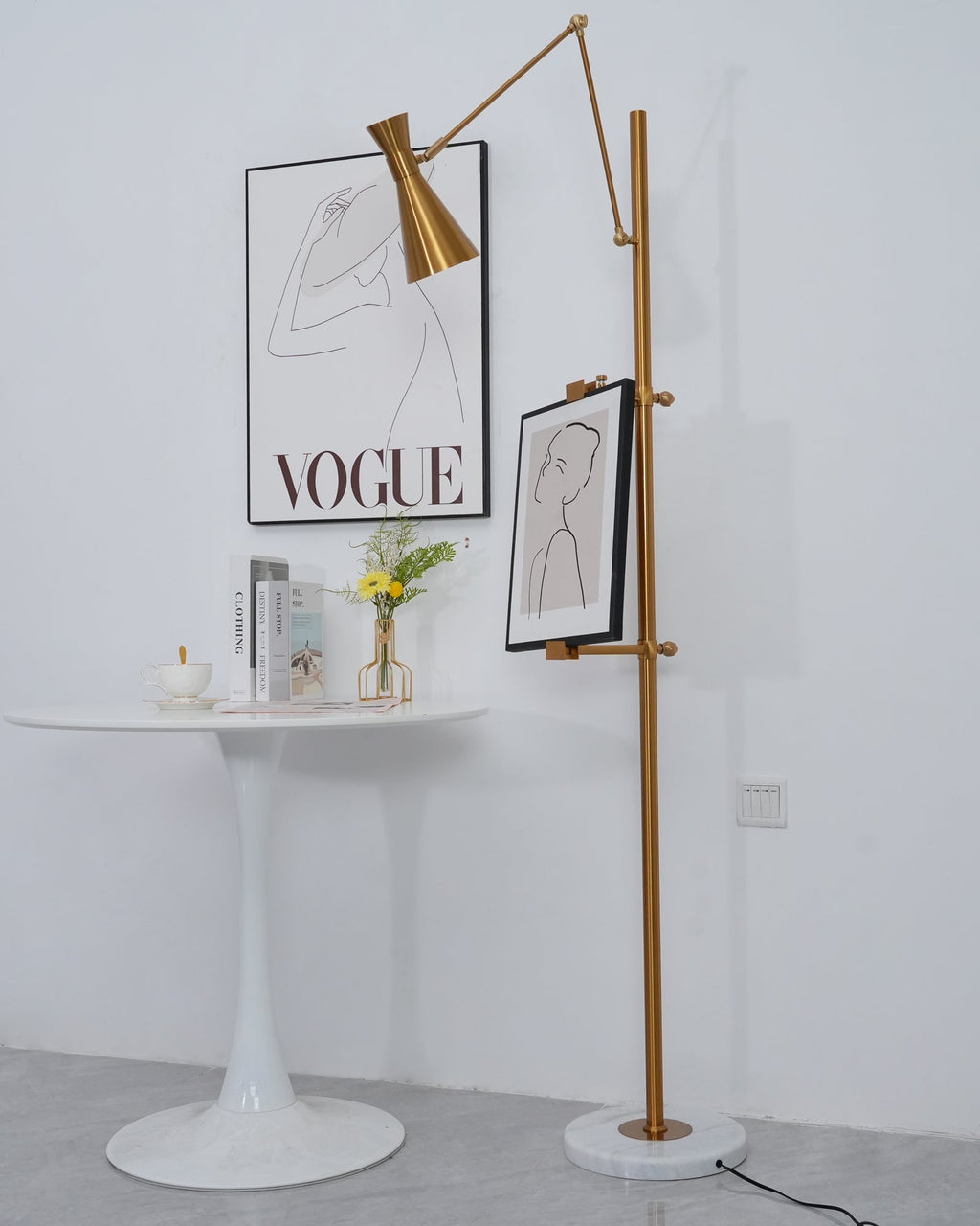 A Clérachic gold floor lamp with a framed drawing beside a round white table, with a printed poster behind and a small vase of flowers.