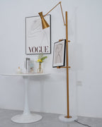 A Clérachic gold floor lamp with a framed drawing beside a round white table, with a printed poster behind and a small vase of flowers.
