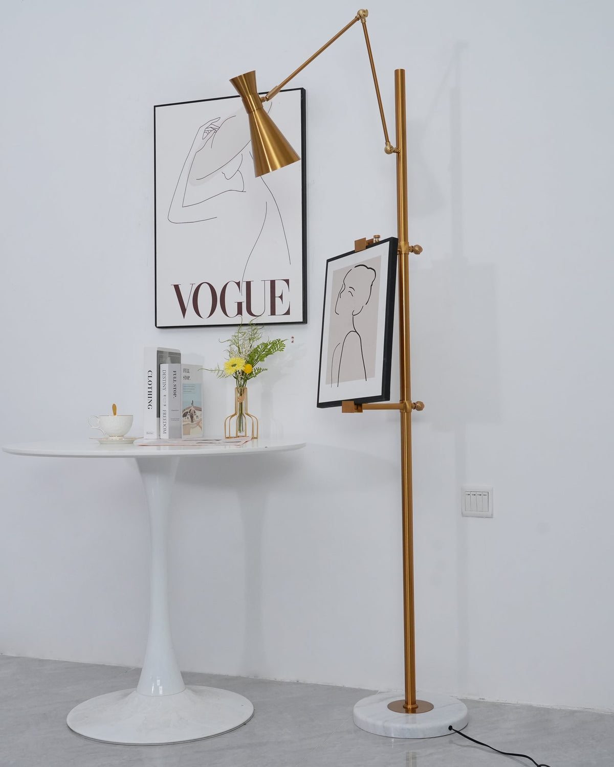 A Clérachic gold floor lamp with a framed drawing beside a round white table, with a printed poster behind and a small vase of flowers.